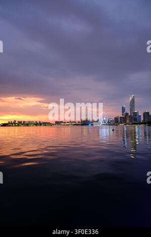 Silhouette calma della città sul lungomare al tramonto con nuvole viola, luci brillanti, riflessi sull'acqua ferma e una ruota panoramica in lontananza. Foto Stock