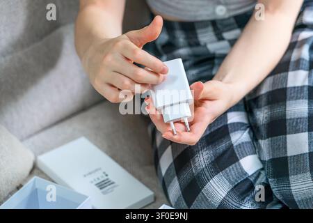 Prima piano di mani che tengono un adattatore di corrente bianco da una scatola di smartphone aperta. Concetto: accessori tecnologici, stile di vita digitale moderno e e-learning Foto Stock