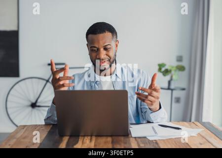 Giovane uomo a un tavolo di legno partecipa a una videoconferenza professionale remota sul suo laptop, gesticolando mentre discute documenti e appunti. Ufficio casalingo casual Foto Stock
