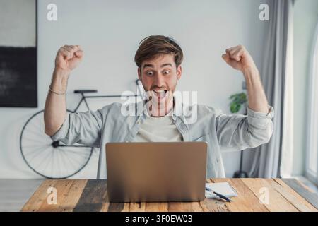 Un giovane alza i pugni in segno di eccitazione mentre guarda il suo laptop in un ufficio domestico luminoso, celebrando il successo online, una vittoria aziendale o un progetto Foto Stock