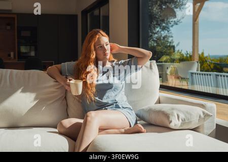Una donna dai capelli rossi è seduta su un divano chiaro tenendo una tazza, si crogiola nella calda luce del sole attraverso grandi finestre e godersi una calma mattina a casa con una rilassata, Foto Stock