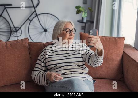 Anziana donna seduta comodamente su un divano del soggiorno, sorride mentre scatta un selfie con il suo smartphone. Momento casuale e rilassato che mostra connessione, te Foto Stock