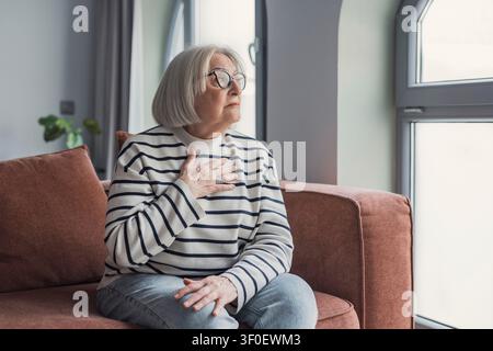 Una donna anziana con occhiali è seduta su un divano vicino a una finestra luminosa, con la mano sul petto e lo sguardo pensieroso verso l'esterno. La scena trasmette riflessione, c Foto Stock