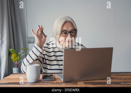 Una donna anziana con gli occhiali si concentra su un laptop a una scrivania di legno, lavorando o partecipando a una videochiamata da casa. Una tazza di caffè, uno smartphone e una pianta Foto Stock