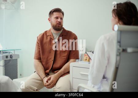 Giovane uomo caucasico che consulta una dottoressa nello studio medico, discutendo sintomi e opzioni di trattamento. Utile per la comunicazione sanitaria, l'assistenza al paziente, il marketing della clinica Foto Stock