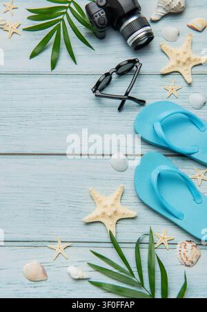 Infradito, macchina fotografica, occhiali da sole, conchiglie e foglie di palma che creano una sistemazione per le vacanze estive su uno sfondo di legno blu Foto Stock