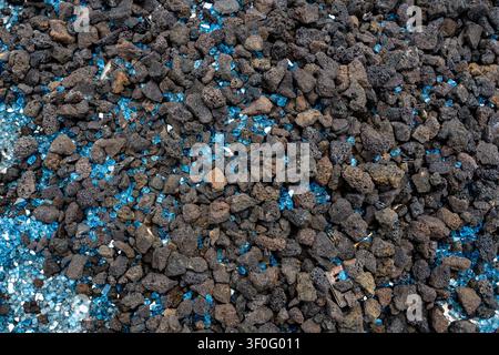 Primo piano di rocce vulcaniche porose marrone scuro mescolate con frammenti di vetro blu brillante, che contrastano la texture naturale ruvida con superfici lisce e riflettenti Foto Stock