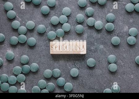 Scatto orizzontale dall'alto di compresse di alghe spirulina e clorella verdi sparse su uno sfondo di pietra grigio scuro intorno a un piccolo blocco di cartello di legno Foto Stock
