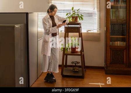 Giovane scienziata in camice da laboratorio bianca in piedi vicino a un supporto per piante in legno con piante in vaso, mentre guarda il suo smartphone in un ufficio caldo e illuminato dal sole con Foto Stock