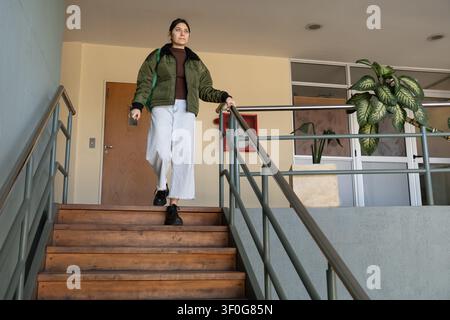 Vista ad angolazione bassa di una giovane donna con giacca bomber verde e zaino che scende scale di legno con ringhiera metallica in un edificio istituzionale luminoso Foto Stock