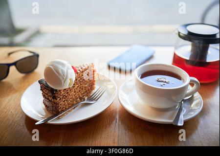 Tè e torta con panna serviti su un tavolo da caffè vicino alla finestra, smartphone e occhiali da sole sullo sfondo, concetto di pausa relax, incontro informale, equilibrio dello stile di vita e relax Foto Stock