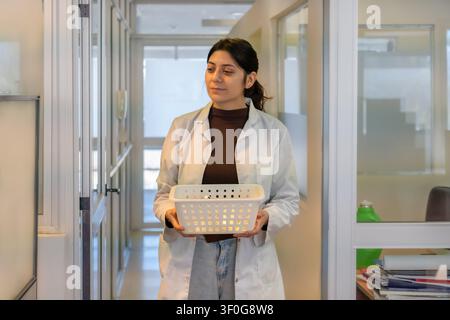 Giovane scienziata in camice da laboratorio bianco che trasporta un vassoio di plastica bianco vuoto attraverso un corridoio istituzionale luminoso con pareti di vetro, guardando t Foto Stock