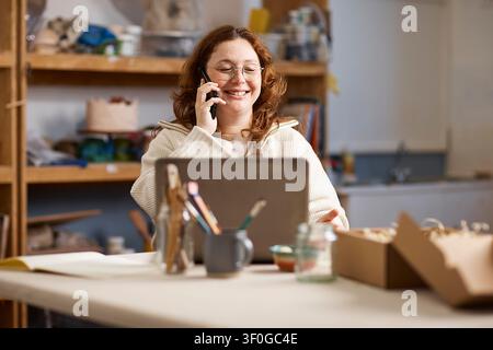 Proprietaria di uno studio di ceramica che sorride al telefono mentre lavora su un laptop e imballa gli ordini Foto Stock