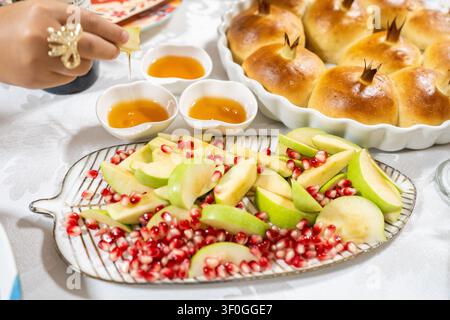 Una persona versa miele su mele affettate su un piatto. I semi di melograno sono sparsi intorno. Cibi dolci segnano la celebrazione di Rosh Hashanah in un ambiente domestico accogliente. Foto Stock