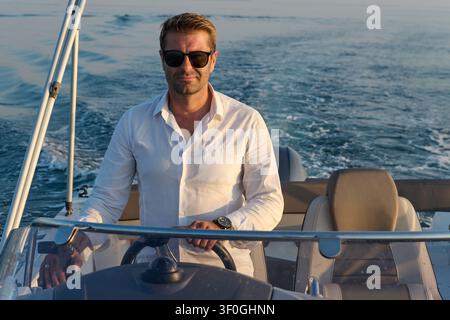 Uomo sicuro di sé alla guida di un motoscafo al tramonto su un mare calmo durante una crociera ricreativa Foto Stock