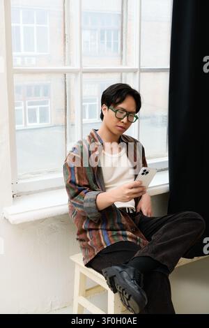 Un giovane uomo asiatico elegante è seduto casualmente vicino a una grande finestra, impegnato con il suo smartphone. Foto Stock