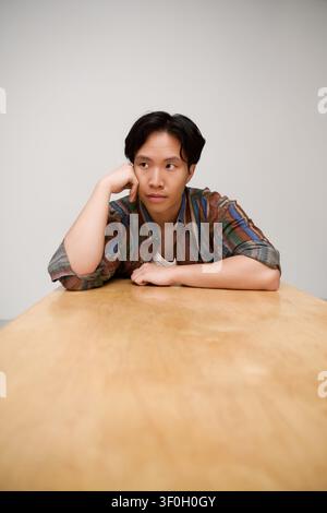 Un giovane uomo elegante siede pensieroso a un tavolo di legno, emanando un'eleganza cool. Foto Stock