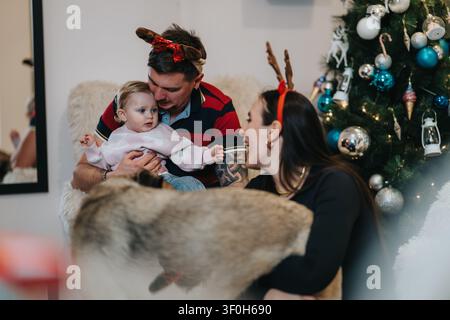 Festa di famiglia a casa con bambino, genitori con corna di renna accanto a un albero di Natale Foto Stock