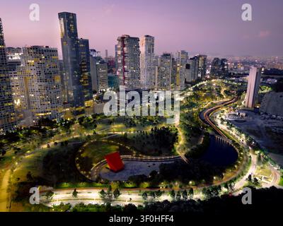 Esposizione aerea con lunga esposizione nell'ora blu che mostra un cielo viola e rosa su un parco urbano illuminato e una skyline moderna con tracce di luce e bagliore della città a S Foto Stock
