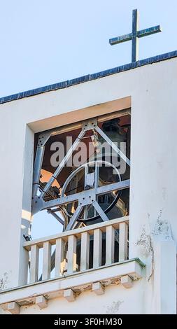 Primo piano di un campanile bianco con una grande campana in metallo e una croce su un cielo blu chiaro, che mostra architettura religiosa e dettagli rustici Foto Stock