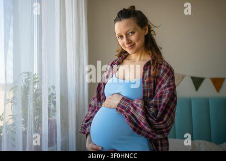 Donna incinta in piedi, tocca delicatamente il pancione e sorride alla telecamera, simboleggiando nuovi inizi, aspettativa e il viaggio della maternità mentre è illuminata dalla luce naturale Foto Stock