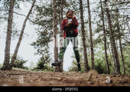 Donna felice che salta in una foresta di pini tenendo dei bastoni su un sentiero escursionistico Foto Stock