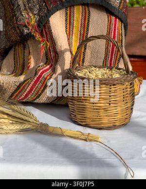 Scatto verticale di un piccolo cesto di vimini intrecciato pieno di grani d'orzo crudi accanto a un mazzo di grano legato e a una sacca tessile cretese a righe tradizionale. Foto Stock