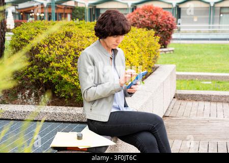 Donna anziana in abiti business casual è seduta all'aperto, concentrata sul suo smartphone. Lo sfondo è caratterizzato dalla vegetazione, creando un ambiente di lavoro sereno. Foto Stock