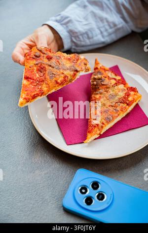 Una persona si gode una pausa informale con due fette di pizza al peperoni croccante su un piatto. Uno smartphone blu giace nelle vicinanze, suggerendo un momento di relax durante Foto Stock