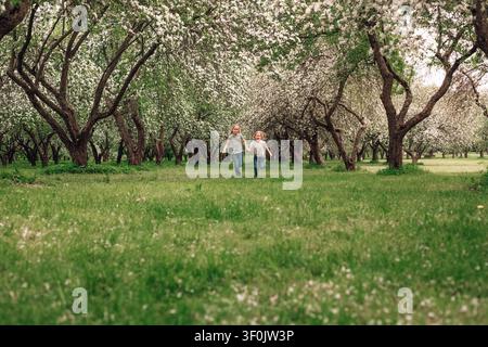 Bambini di una famiglia felice corrono mano nella mano attraverso un frutteto in fiore primaverile, circondati da alberi in fiore e erba verde fresca, catturando momenti gioiosi all'aperto Foto Stock