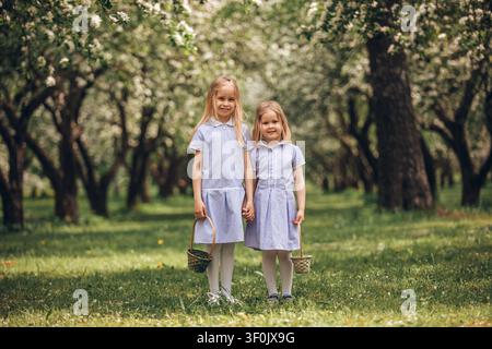 Due sorelle sorridenti si tengono per mano in un frutteto in fiore in primavera, tenendo piccoli cestini sull'erba verde, godendo della luce calda del giorno e di una famiglia felice Foto Stock