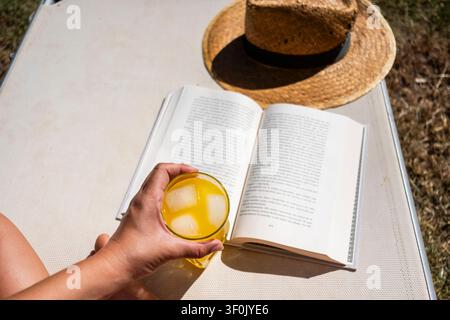 Una scena rinfrescante di relax estivo con un libro aperto e una bevanda fredda con cubetti di ghiaccio in una giornata di sole. Un cappello di paglia riposa nelle vicinanze, completando Foto Stock