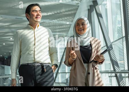 Due colleghi di lavoro asiatici camminano e chiacchierano in un corridoio d'ufficio moderno, sorridendo mentre discutono di lavoro. Donna con hijab tiene un tablet alla luce naturale brillante Foto Stock