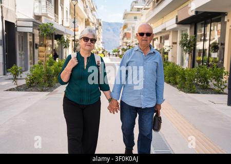 Coppia di anziani che si tengono per mano mentre camminano lungo una strada pedonale soleggiata a Corinto, Grecia, godendosi il turismo culturale mediterraneo in un Ci storico Foto Stock
