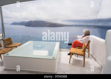 Donna anziana seduta su una terrazza di un hotel di lusso a Santorini, Grecia, godendosi una tranquilla vista sul mare sopra la caldera con vasca idromassaggio privata, sedie moderne, un Foto Stock