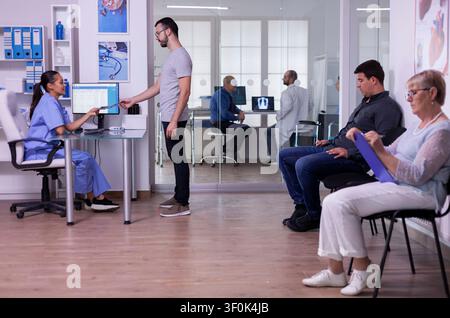 Pazienti che compilano moduli medici in una sala d'attesa affollata, giovane uomo che fa una radiografia a un'infermiera, fissando un nuovo appuntamento. Medico specialista che consulta un anziano nella sala visita. Foto Stock