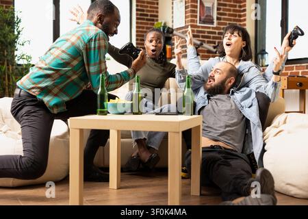 Gruppo diversificato ed entusiasta di amici che esultano al chiuso durante un ritrovo del fine settimana. Gioiosi giocatori multietnici celebrano la vittoria con mani alzate, risate e legame su momenti divertenti di videogiochi. Foto Stock