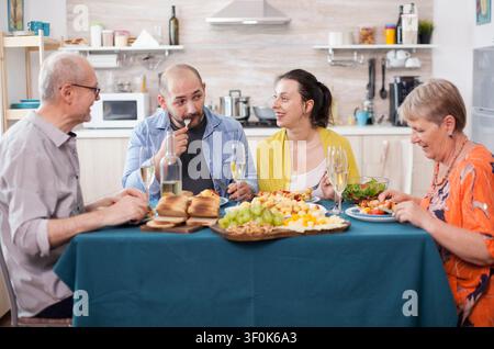 La figlia sorride al padre durante un pranzo familiare in cucina, con patate arrosto e cibo condiviso a un tavolo da pranzo felice. Foto Stock