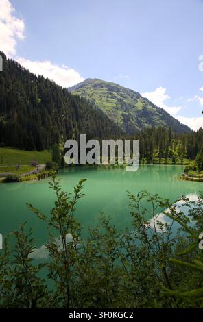 Lago alpino turchese che riflette una foresta sempreverde e pendici montuose, con una piccola centrale idroelettrica sulla riva sotto la luce naturale del giorno in Austri Foto Stock