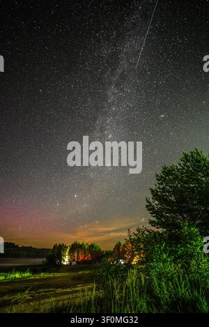 La Via Lattea si estende verticalmente attraverso un cielo notturno denso di stelle sopra una riva tranquilla di un lago boschivo nella campagna russa. Foto Stock