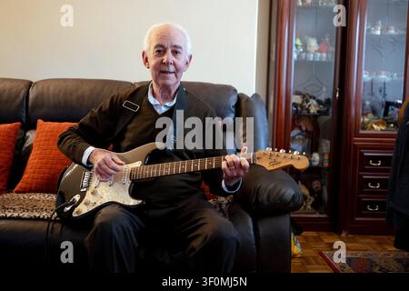 Uomo anziano seduto su un divano che suona una chitarra elettrica, godendosi la musica a casa Foto Stock