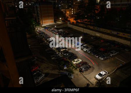 Una vista notturna ad angolo elevato di un parcheggio residenziale affollato. L'esposizione lunga cattura vivaci scie di luce rosse da un veicolo in movimento, contrasto Foto Stock