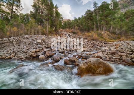 Un ruscello di montagna dal flusso veloce cade a cascata su un ampio ventaglio alluviale roccioso prima di confluire in un fiume turchese più grande. Foto Stock