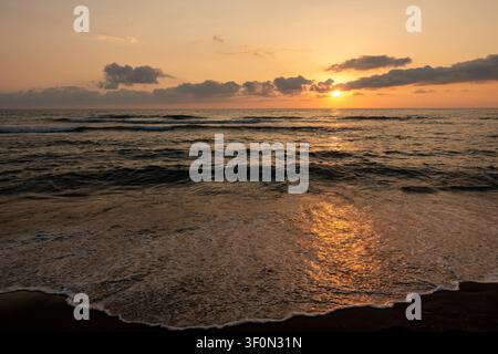La luce calda del tramonto bagna una spiaggia sabbiosa mentre dolci onde oceaniche si infrangono, proiettando un riflesso dorato vivido sul sabbia bagnata e sulla schiuma. Foto Stock