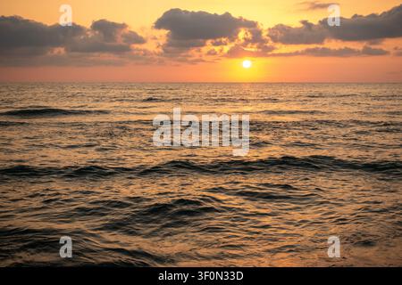 Un ampio paesaggio marino cattura il sole che scende verso l'orizzonte sopra le onde dell'oceano che si arrotolano dolcemente. Foto Stock