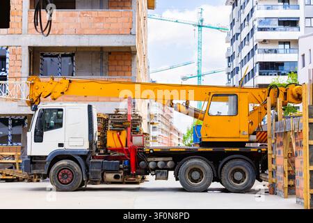 Veicolo gru camion giallo parcheggiato nel cantiere di costruzione di un edificio residenziale con gru torrelo in sottofondo Foto Stock