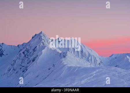 Cime innevate si stagliano contro un cielo vivace di alpenglow rosa e viola durante la transizione dell'ora blu. Foto Stock
