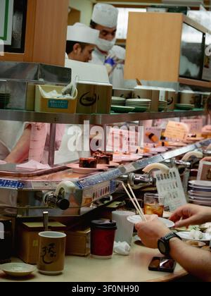 Chef che preparano il cibo dietro il nastro trasportatore di un ristorante sushi kaiten-zushi a Shibuya, Tokyo, Giappone, mentre un cliente mangia con le bacchette Foto Stock
