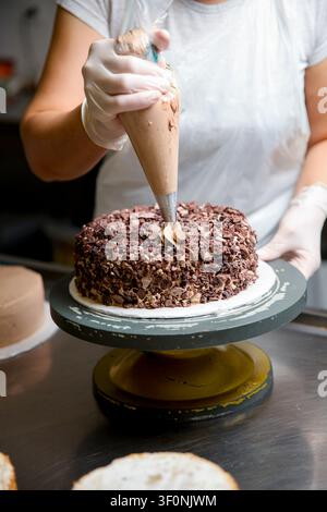 Un panettiere lavora in una cucina. Usano una sac à poche per aggiungere glassa a una torta al cioccolato. Altri strati di torta sono posizionati nelle vicinanze su un tavolo. Foto Stock