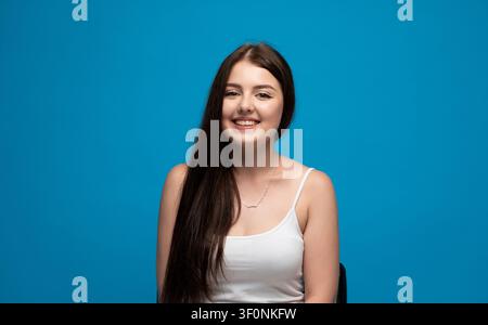 Giovane donna che sorride ampiamente, mostra felicità ed emozioni positive su uno sfondo blu vibrante in studio Foto Stock